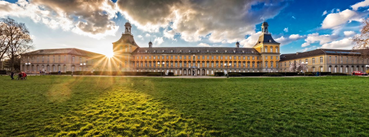 Blick von der Parkanlage Hofgarten auf die gelbe Fassade des Kurfürstlichen Schlosses Bonn, dem Hauptgebäude der Rheinischen Friedrich-Wilhelms-Universität. Über dem Gebäude strahlt die Sonne.