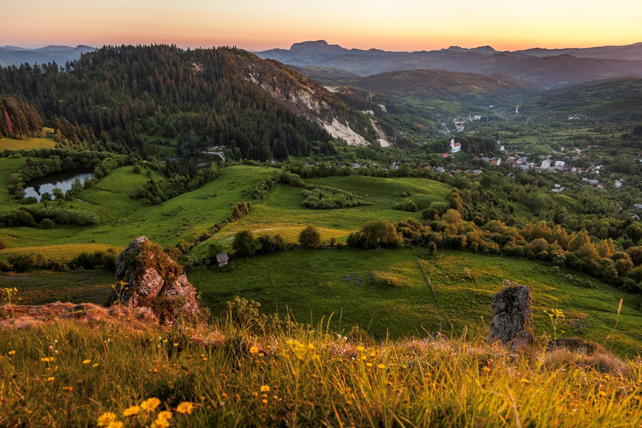 Eine Berglandschaft mit Wiesen und Wäldern bei Roșia Montană in Rumänien ist in orangefarbenes Abendlicht getaucht.