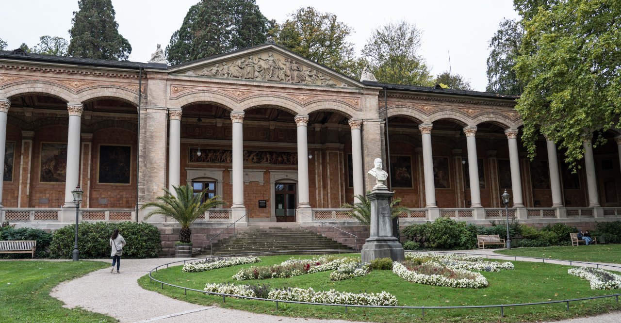 Baden-Badens Trinkhalle von außen, ein prächtiges Gebäude mit Säulengang, davor ein Garten mit einem Rondell aus Blumen. In der Mitte eine Büste des Kaisers Wilhelm I.