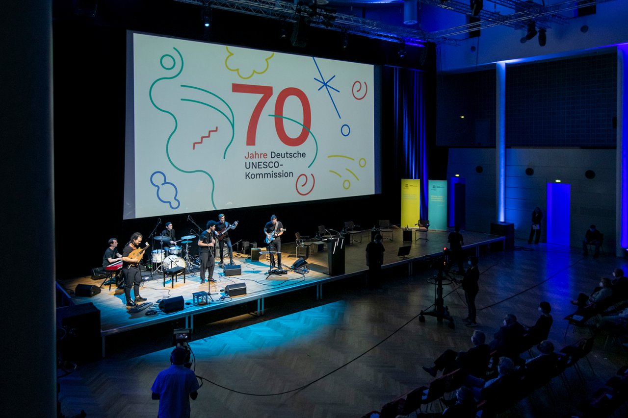 Eine Bühne auf der eine Band im Scheinwerferlicht musiziert, dahinter eine große Grafik mit "70 Jahre Deutsche UNESCO-Kommission"