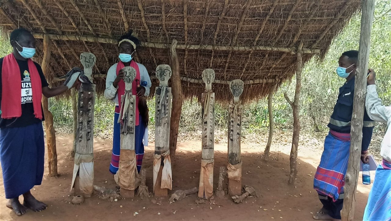 mehrere Personen unter einem einfachen Unterstand mit 5 Holzstatuen