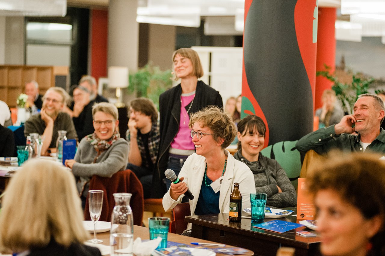 junge Menschen sitzen an einem Tisch, essen und unterhalten sich. Im Hintergrund sind weitere Tische mit Menschen zu erkennen.