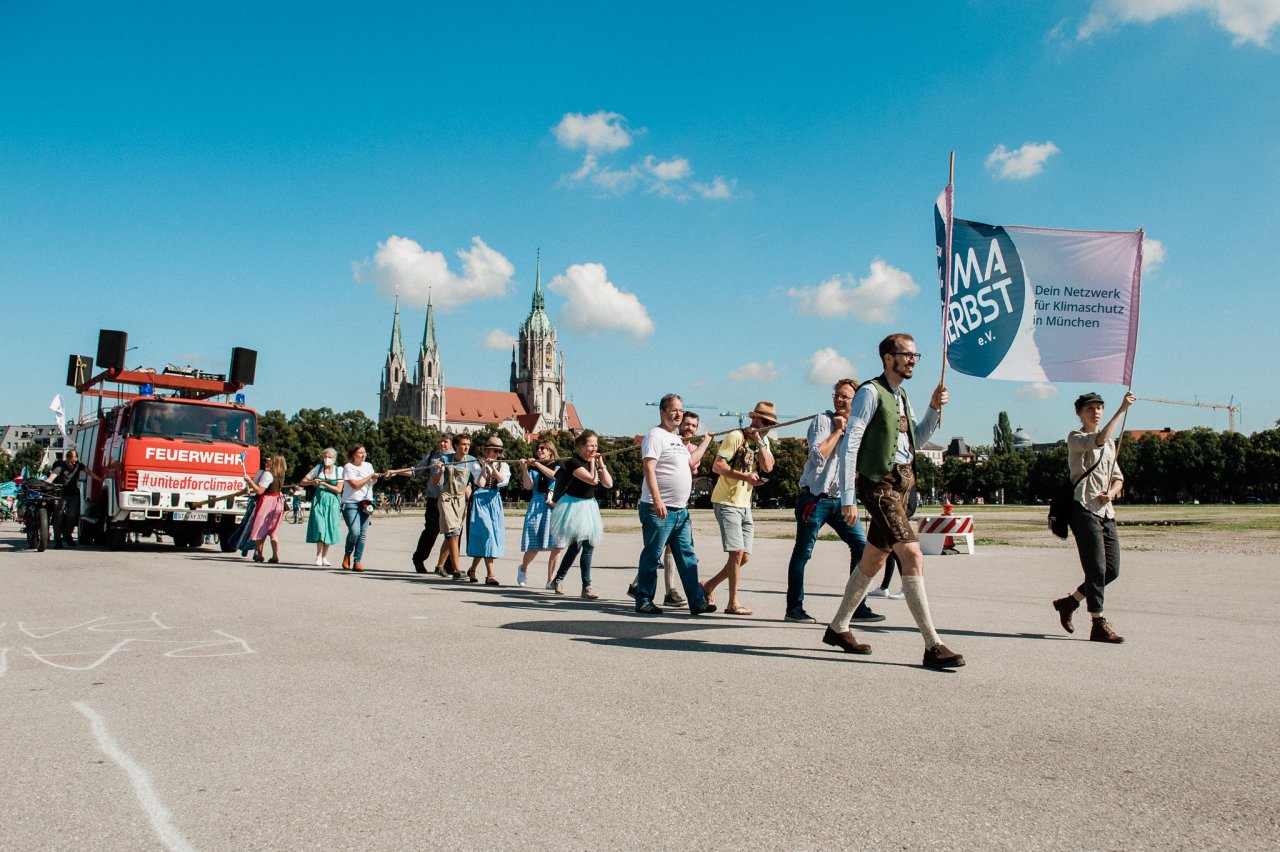 Der Demonstrationszug des "Wiesneinzug der Klimaheld*innen 2021" ist zu sehen: Menschen, die ein Banner des "Netzwerk Klimaherbst e.V." hochalten und ein ehemaliges Feuerwehr Auto von Hand ziehen