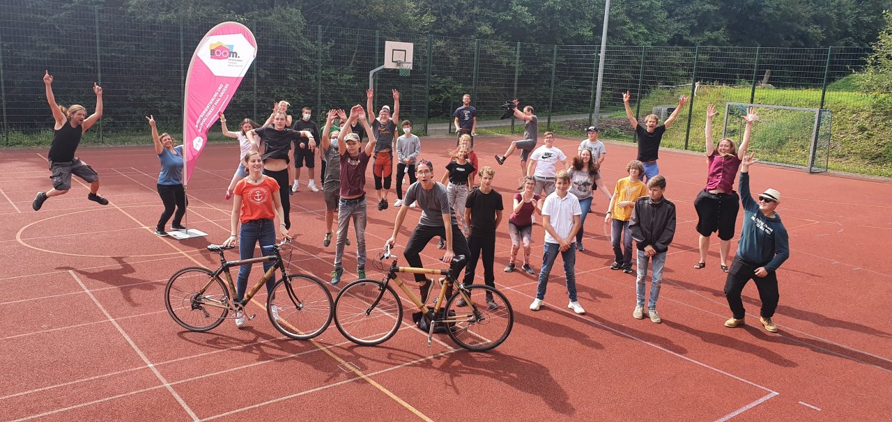 Eine Gruppe junger Menschen freut sich auf einem Sportplatz über den gelungenen Abschluss eines BOOM Feriencamps. Mit dabei sind zwei im Camp erstellte Bambus-Fahrräder