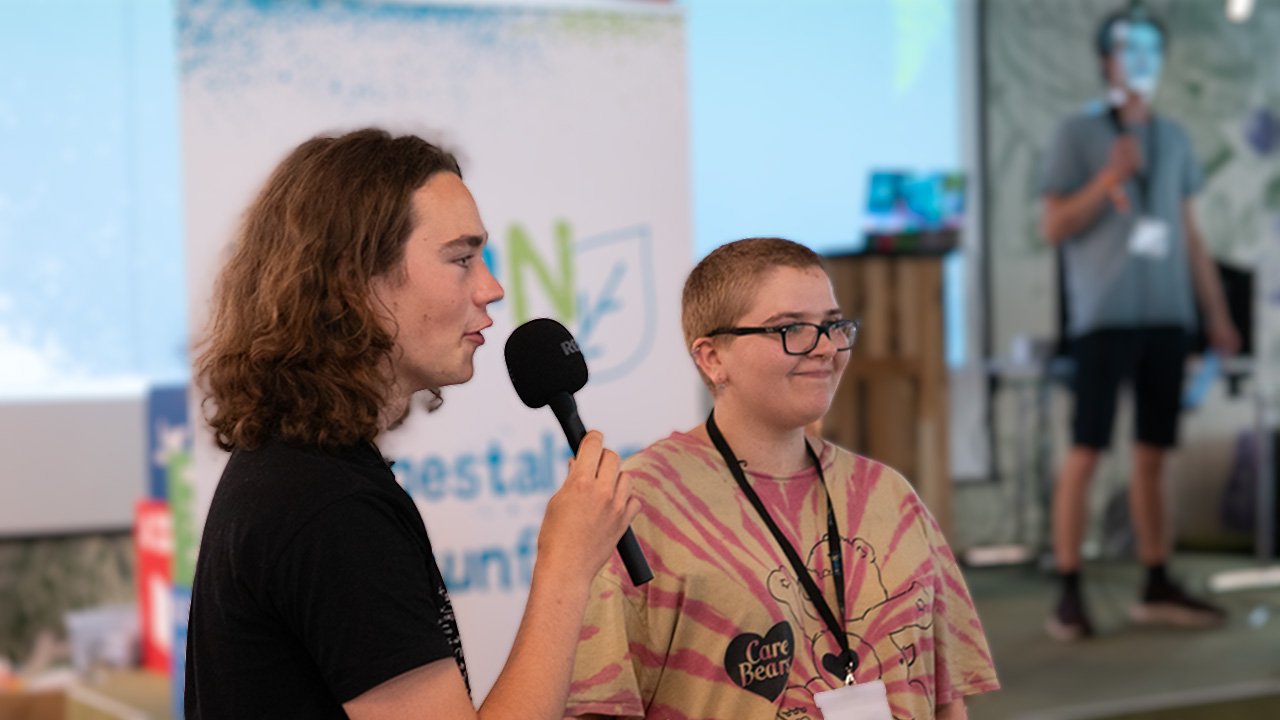 Zwei junge Menschen stehen auf der Jugendkonferenz youcoN vor einer Bühne. Eine der Personen spricht in ein Mikrofon. Im Hintergrund steht ein weiterer junger Mensch auf der Bühne.