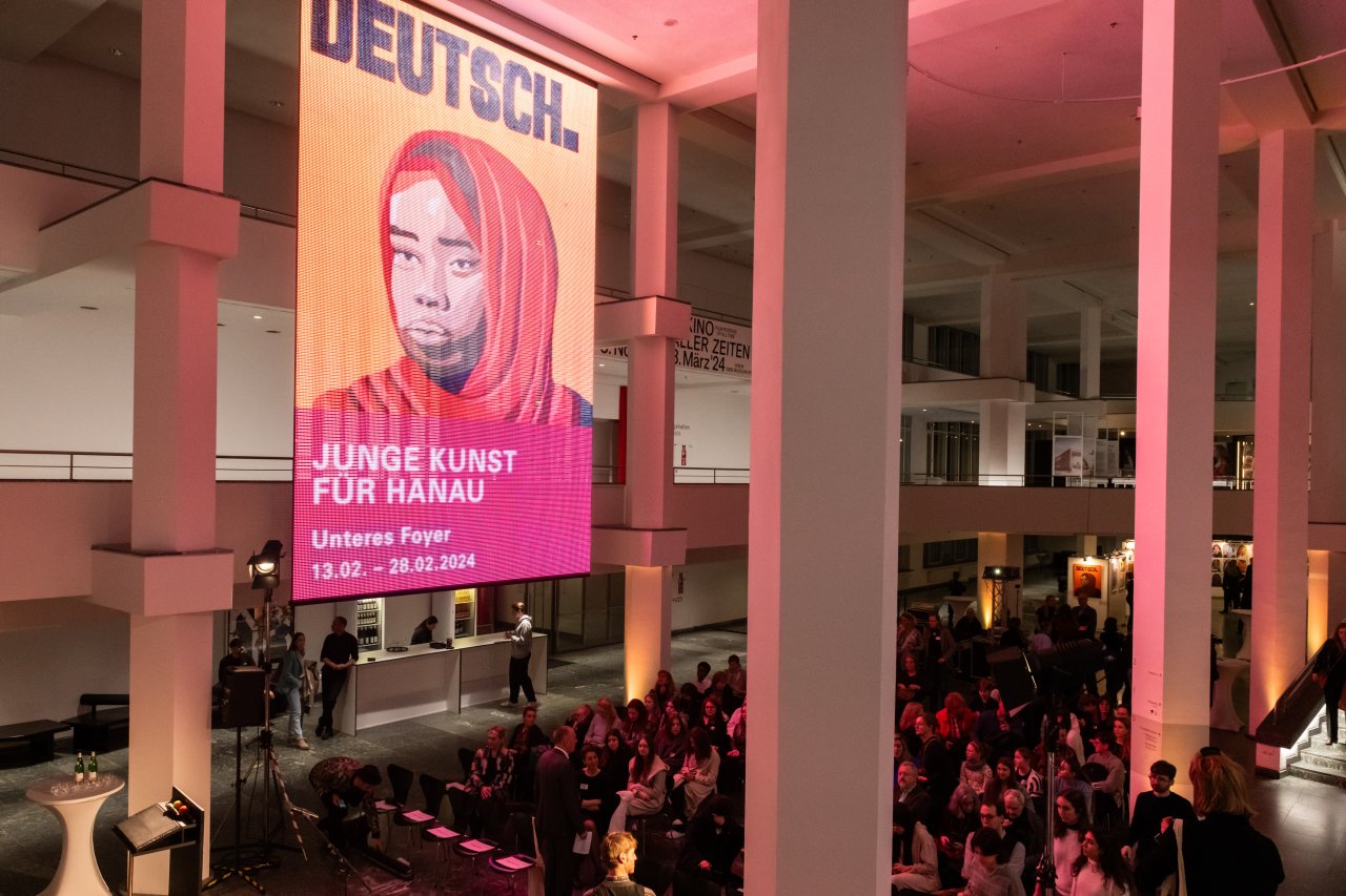 Ein Asustellungsbanner mit dem Kunstwerk einer Schülerin, welches eine junge Frau mit ernstem Blick und der Überschrift "Deutsch." zeigt, hängt großformatig hoch oben in einem Veranstaltungssaal, in dem sich viele Besucherinnen und Besucher aufhalten.