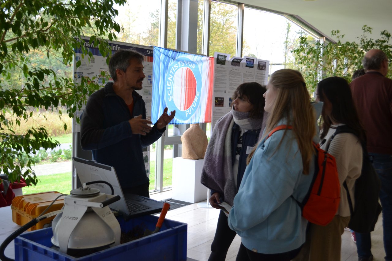 Ein Wissenschftler im Gespräch mit drei Oberstufenschülerinnen in Konferenzumgebung. Im Hintergrund ein Banner von Scientists for Future