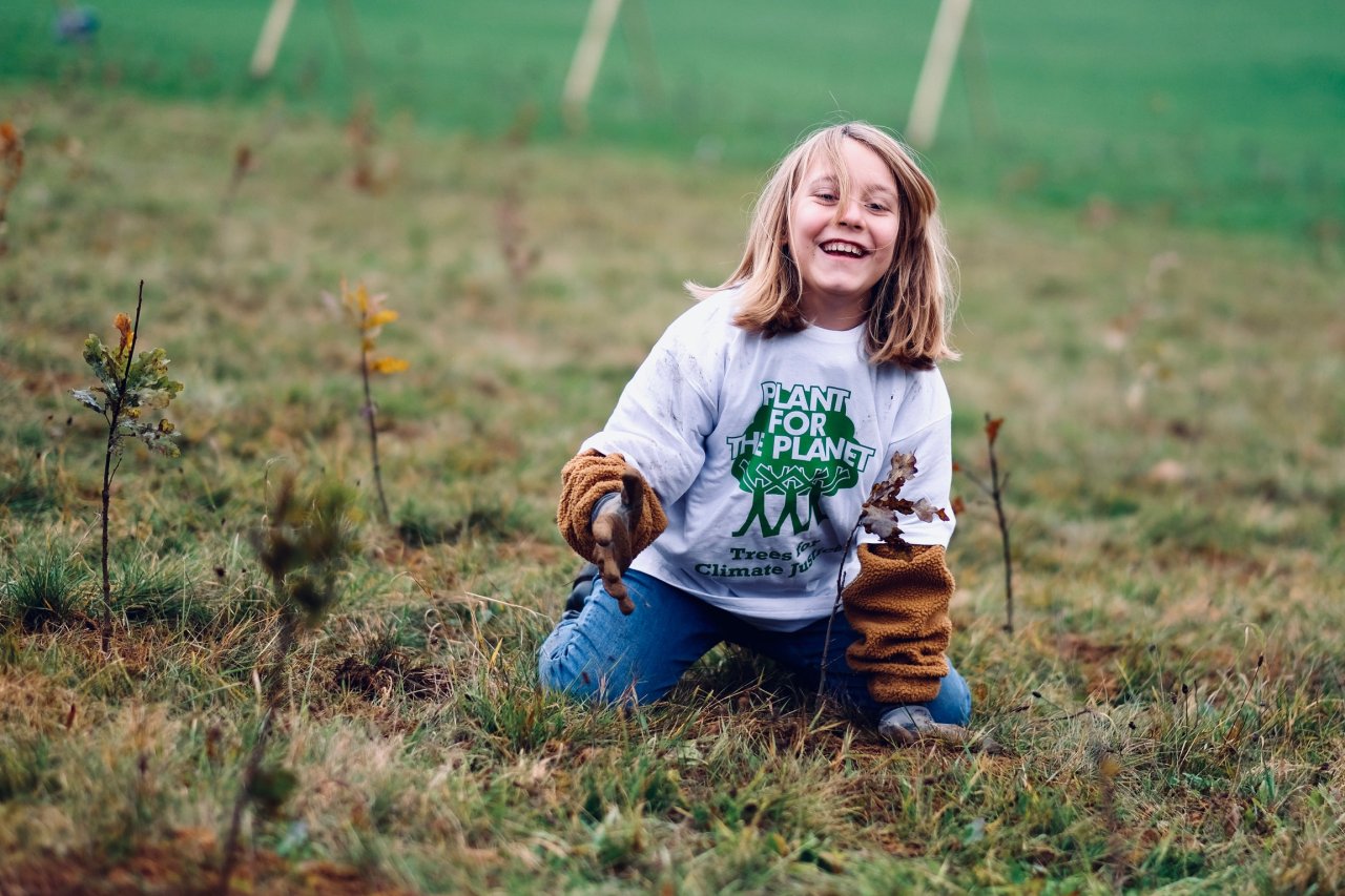 Ein Kind hockt auf einer Grünfläche, pflanzt einen Baum und lacht in Kamera.