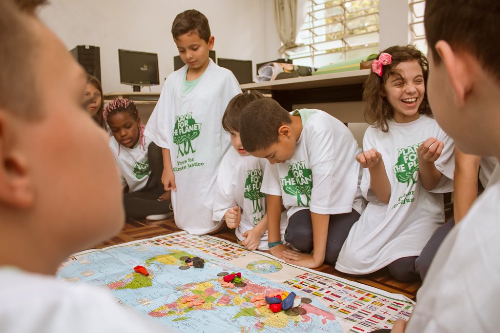 Kinder sitzen auf einer Akademie um eine Weltkarte, auf der Luftballons, Menschenfiguren und Münzen liegen.