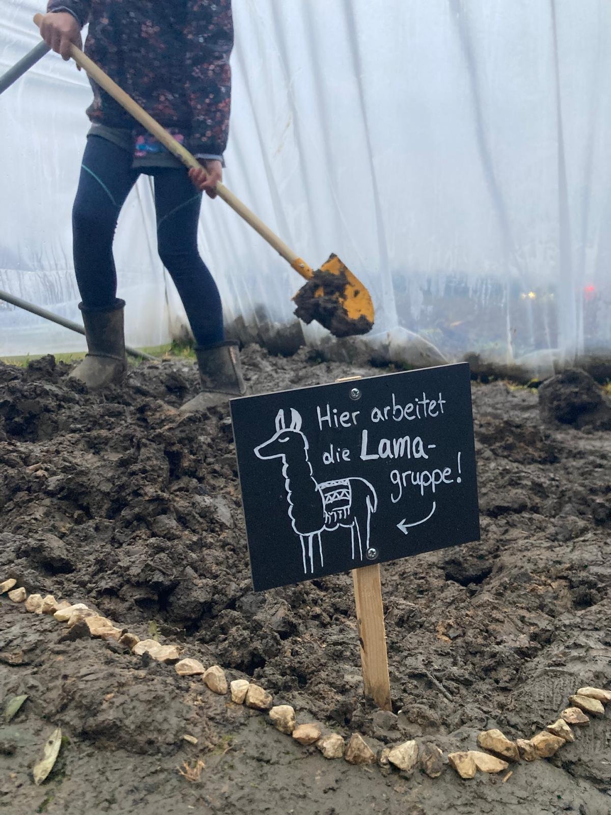 Im Hintergrund gräbt ein Kind im Folientunnel die Erde um. Im Vordergrund ist ein handgeschriebenes Schild, auf welchem steht: Hier arbeitet die Lama-Gruppe!