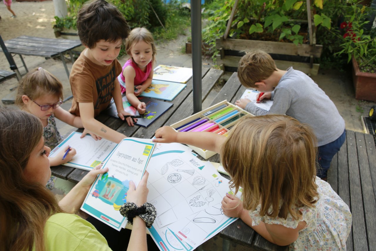 Fünf kleine Kinder im Kita-Alter sitzen um einen Tisch, auf dem Bildungsmaterialien und Stifte verteilt liegen. Sie malen Bilder bunt aus. Eine erwachsene Person sitzt im Vordergrund.