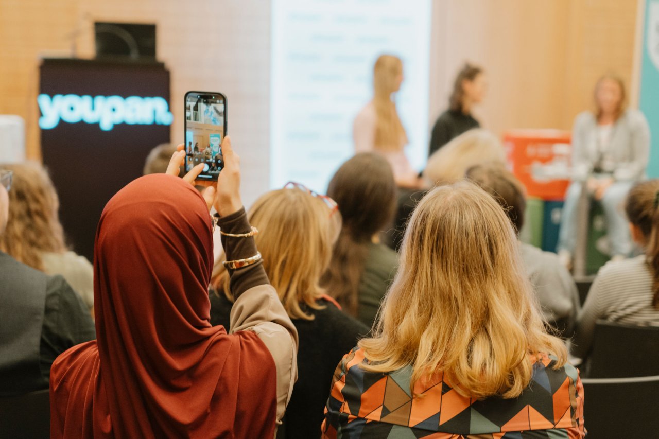 Person mit Hijab sitzt im Publikum und macht ein Handy-Foto von der Bühne vor ihr. Auf der Bühne sitzen zwei youpan-Mitglieder sowie eine erwachsene Person. Links daneben wird das youpan-Logo auf einem Rednerpult angezeigt.