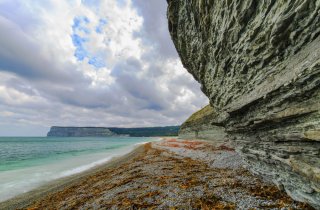 Küstenaufnahme mit Felsen auf der rechten Seite, einem kleinen Strandstück in der Mitte und links das smaragdfarbene Meer.