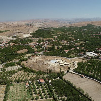 Blick über eine hügelige Landschaft mit bepflanzten Hängen und einem Hügel mit den Überresten der Siedlungen von Arslan Tepe.