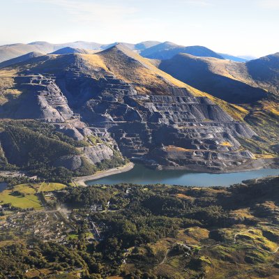 Blick über eine weite Berglandschaft mit Hängen die zum Teil grün bewachsen, zum Teil aus schwarzem Schiefer sind. Im Tal liegt ein Gewässer.