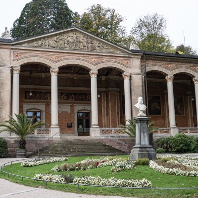 Baden-Badens Trinkhalle von außen, ein prächtiges Gebäude mit Säulengang, davor ein Garten mit einem Rondell aus Blumen. In der Mitte eine Büste des Kaisers Wilhelm I.